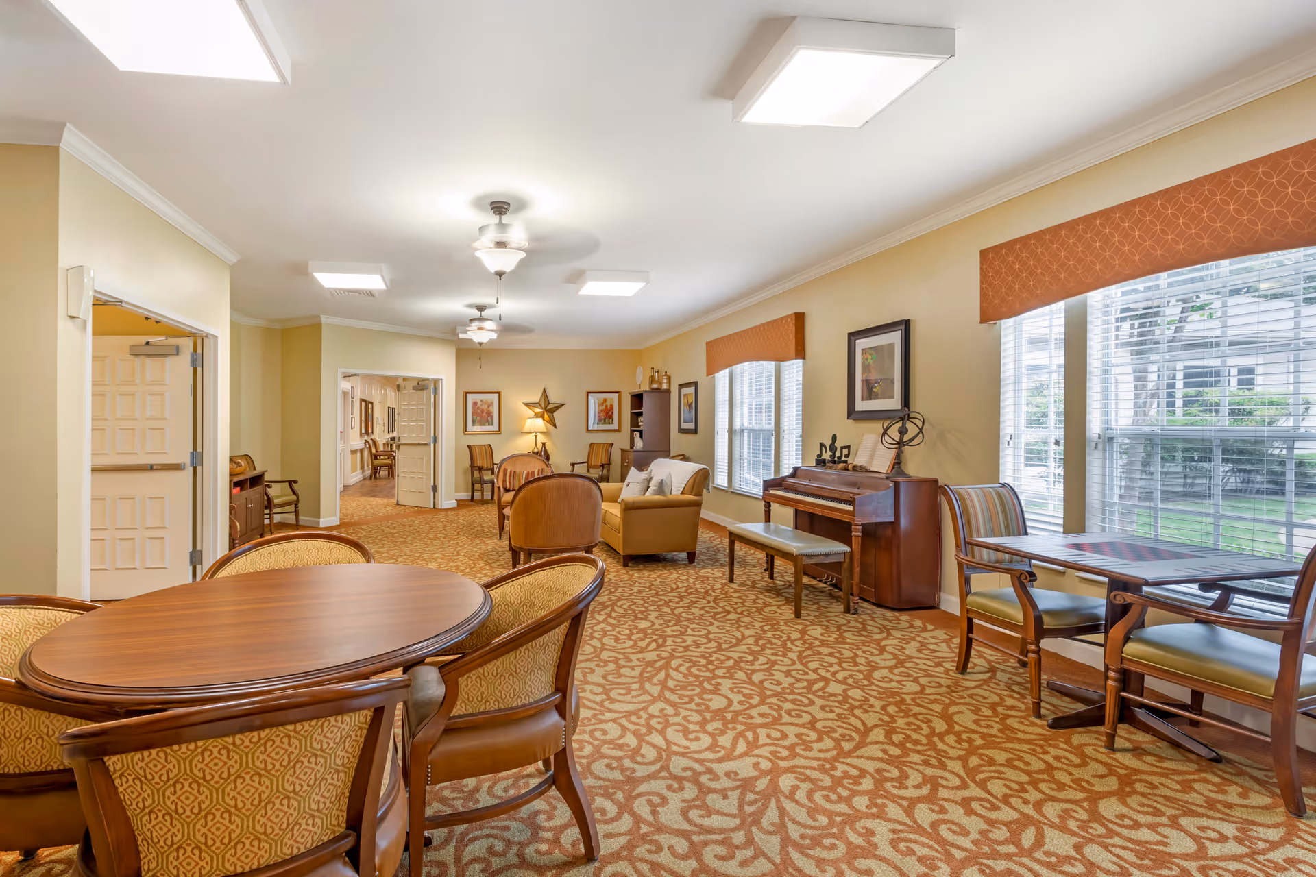 Bright carpeted common room with round dining tables, chairs, sofas, and a piano beside large windows.