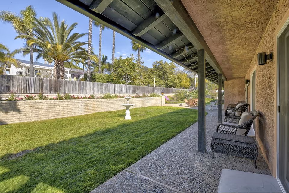 A covered patio area with wicker chairs and cushions along the side of a building, overlooking a well-maintained grassy yard with a decorative birdbath and palm trees in the background under a clear blue sky.