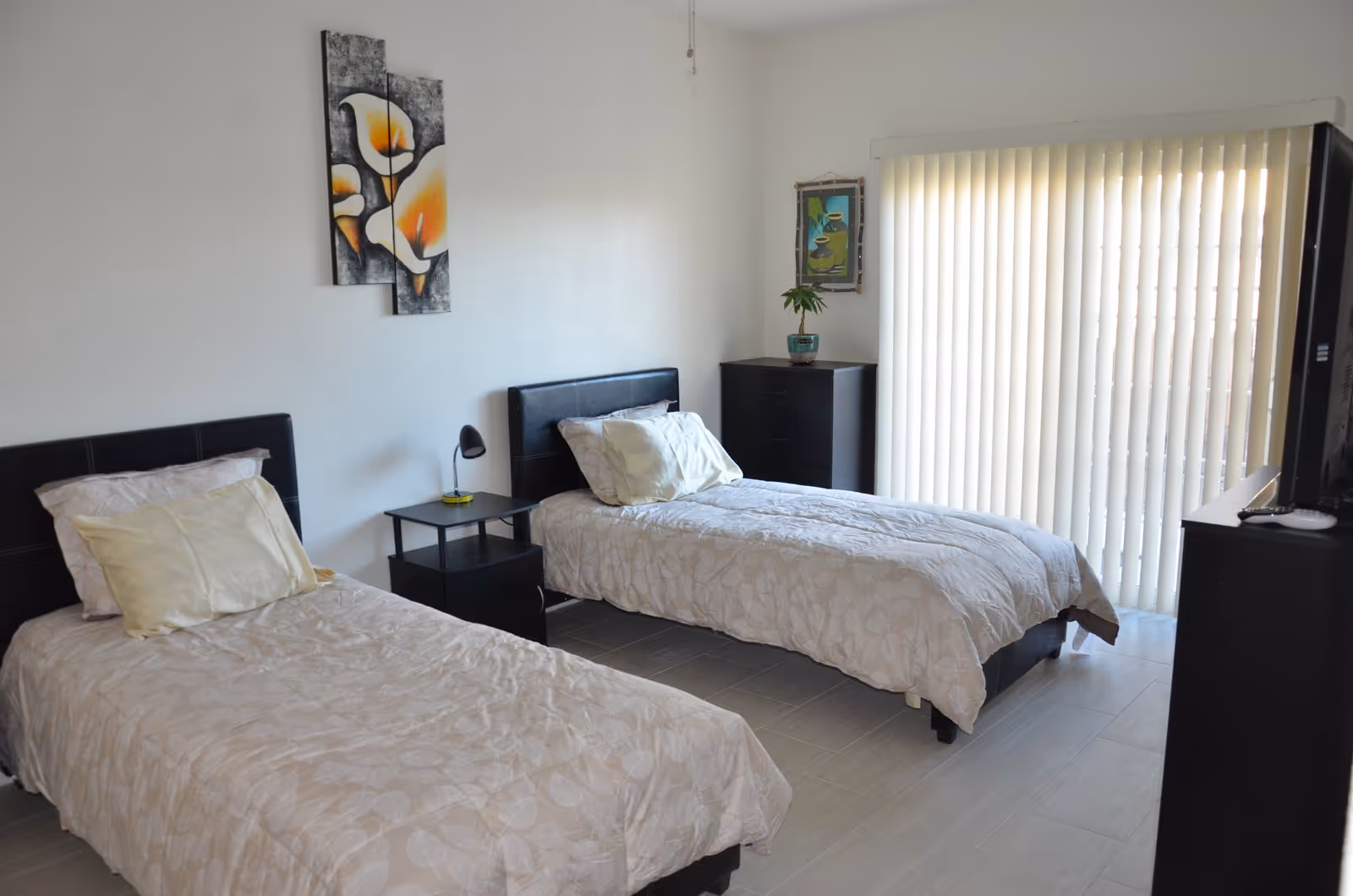 A bedroom with two single beds, each with light-colored bedding and pillows. Between the beds is a small black nightstand with a lamp. On the wall above one bed is a three-panel painting of orange and white flowers. There is a black dresser with a small potted plant and a framed picture on the wall above it. Vertical blinds cover a large window or sliding door, and a television is visible on the right side of the room.