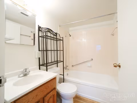 A small bathroom with a white sink and wooden cabinet, a toilet, and a bathtub with a shower curtain rod. There is a metal shelving unit above the toilet and a mirror above the sink. The walls and tiles are white, and the floor has a wood-like finish.