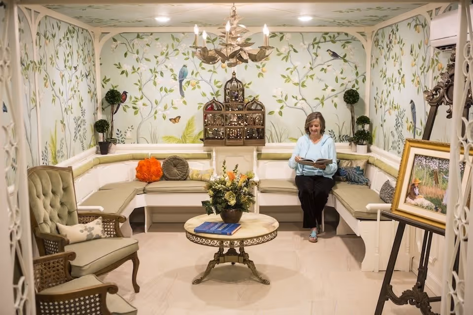 A cozy sitting area with a woman reading a book on a cushioned bench that wraps around the room. The walls are decorated with a mural of leafy branches and birds. There is a round table with a flower arrangement and books in the center, a green upholstered armchair, potted plants, and a framed painting on an easel.