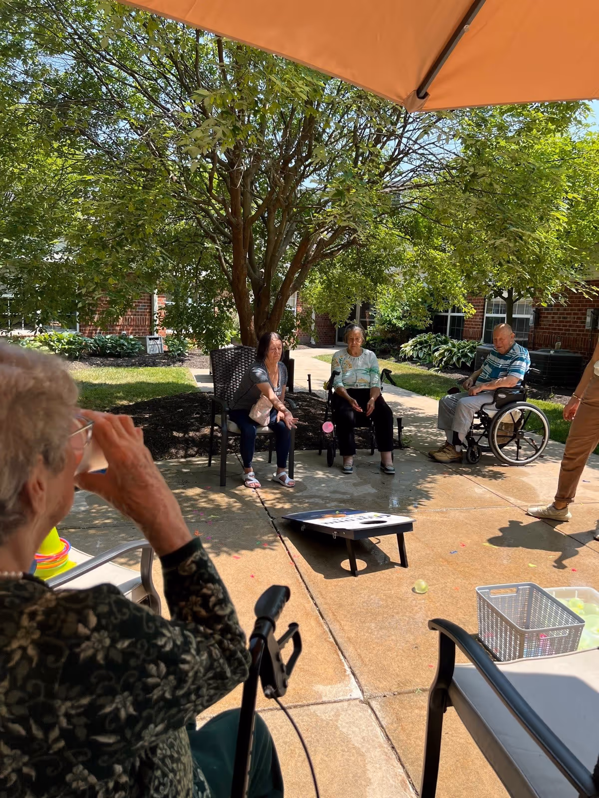 A group of elderly people sitting outdoors on a sunny day, playing a game of cornhole on a paved patio area with trees and greenery in the background. One person is in a wheelchair, and others are seated on chairs. An orange umbrella provides shade overhead.