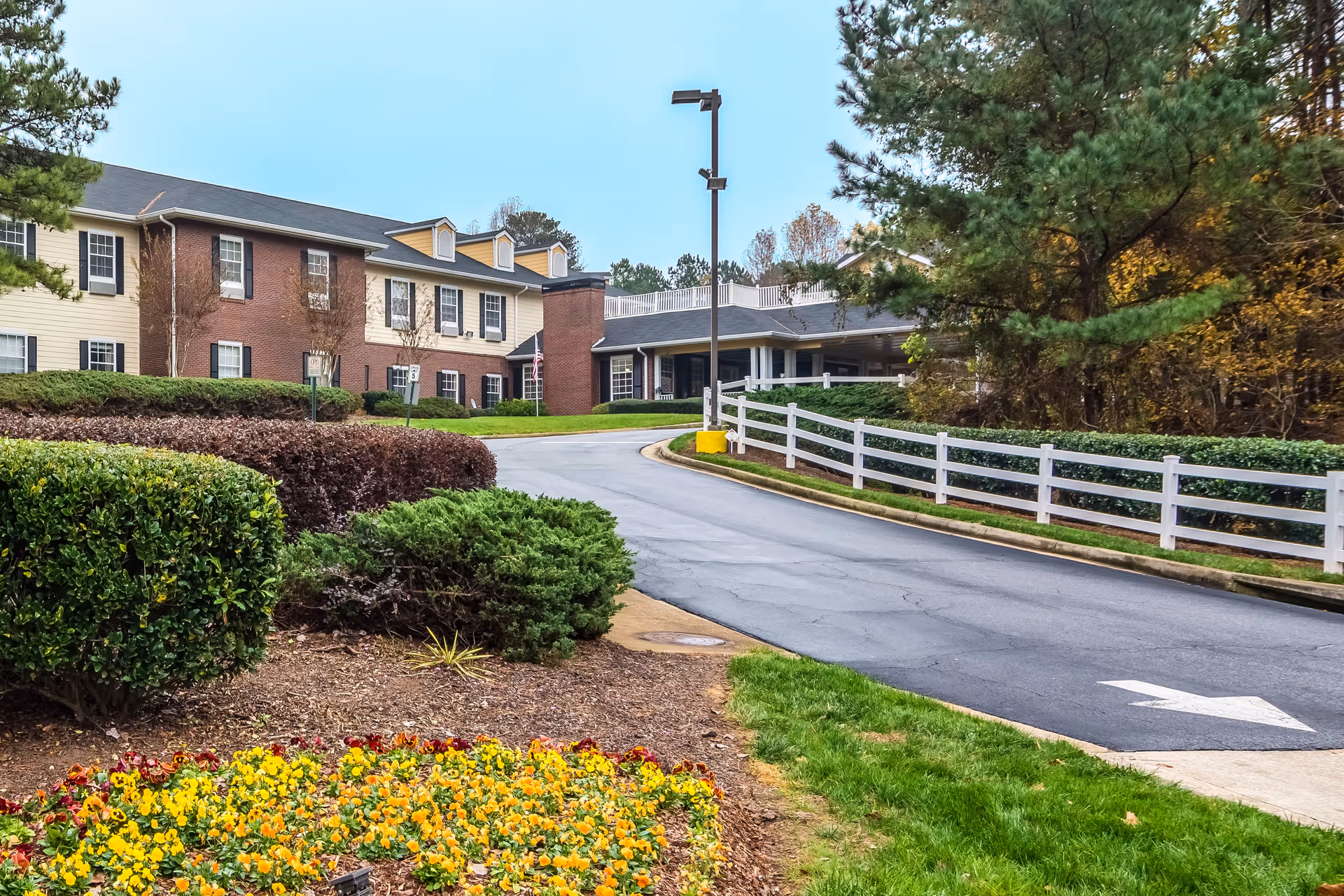 Driveway entrance to The Retreat at Cascade showing a two-story brick-and-siding building, landscaped flowerbeds, and a white fence.