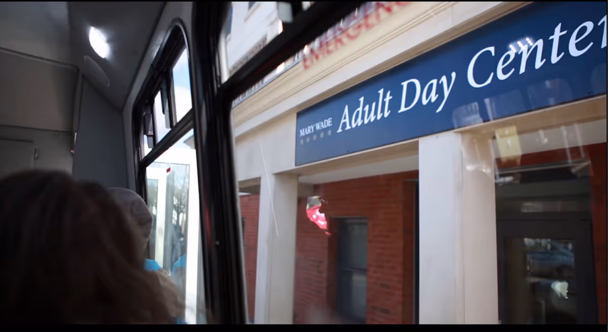 View from inside a vehicle looking out the window at the Mary Wade Adult Day Center building with a blue sign above the entrance.