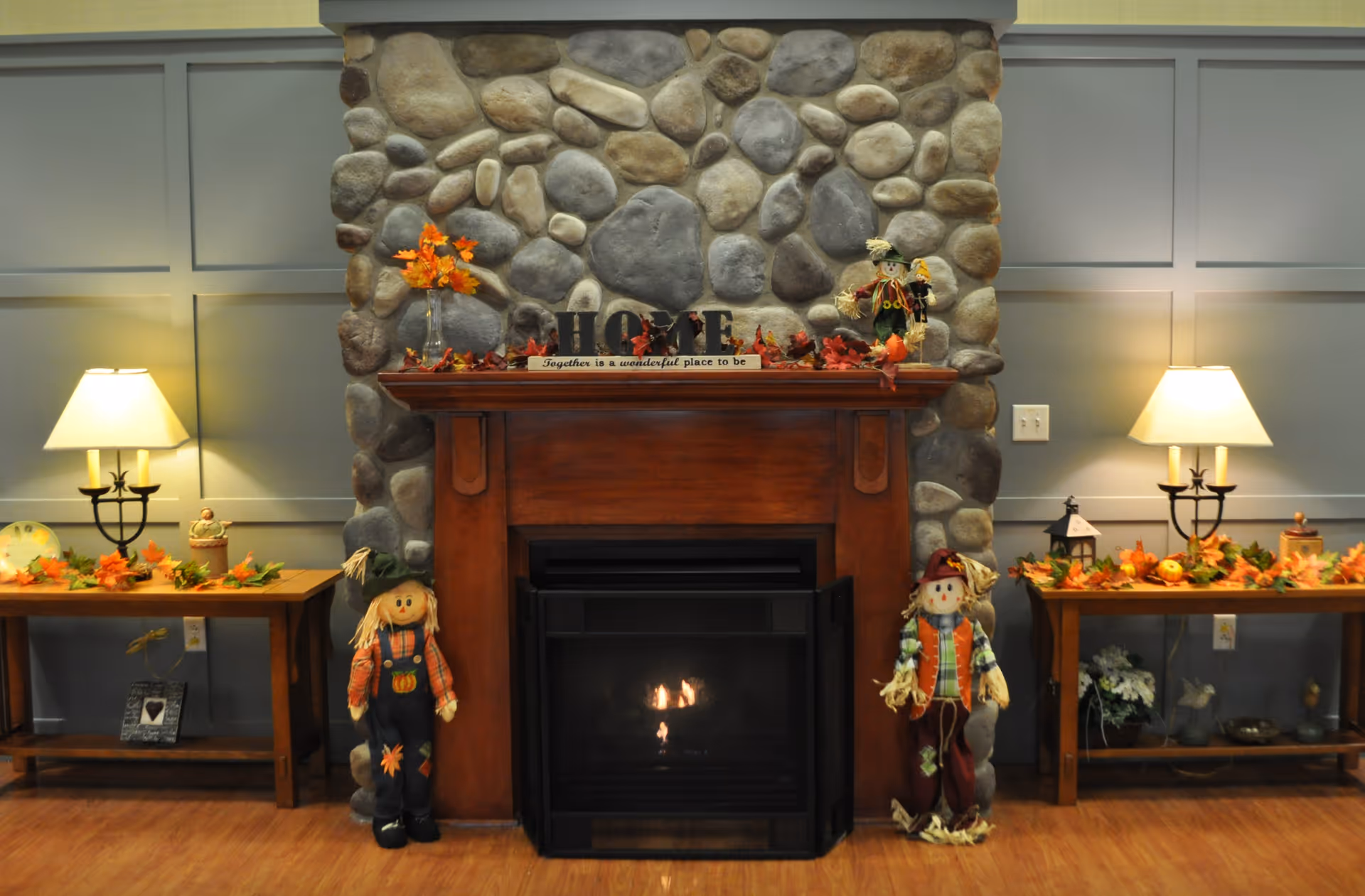 Cozy stone fireplace with a wooden mantel decorated for fall, flanked by lamps, side tables and scarecrow figures.