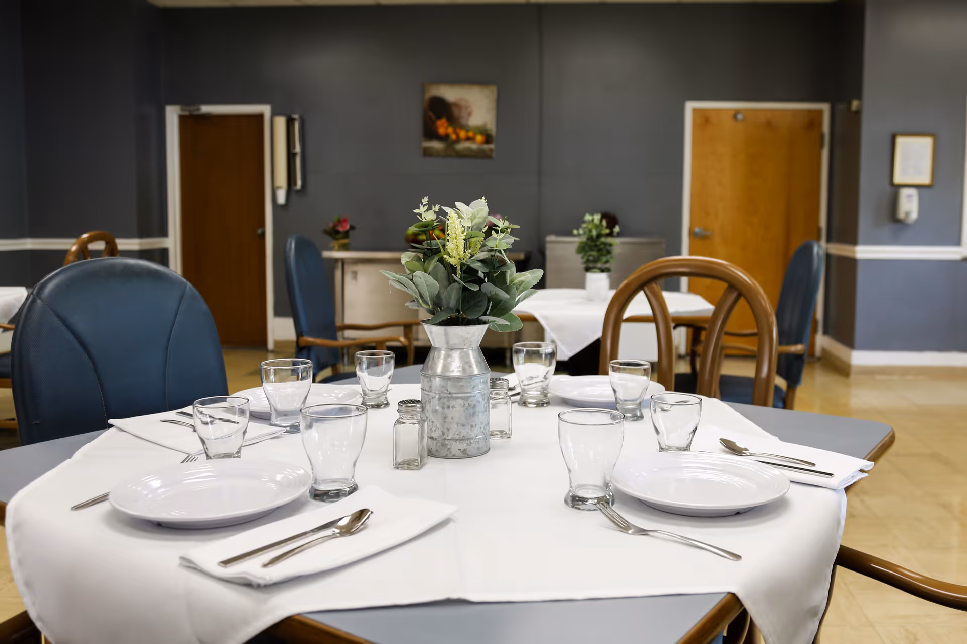A dining room table set for six with white plates, drinking glasses, silverware on white napkins, and a metal vase with green foliage as a centerpiece. The room has blue-gray walls, wooden doors, and additional tables with similar settings in the background.