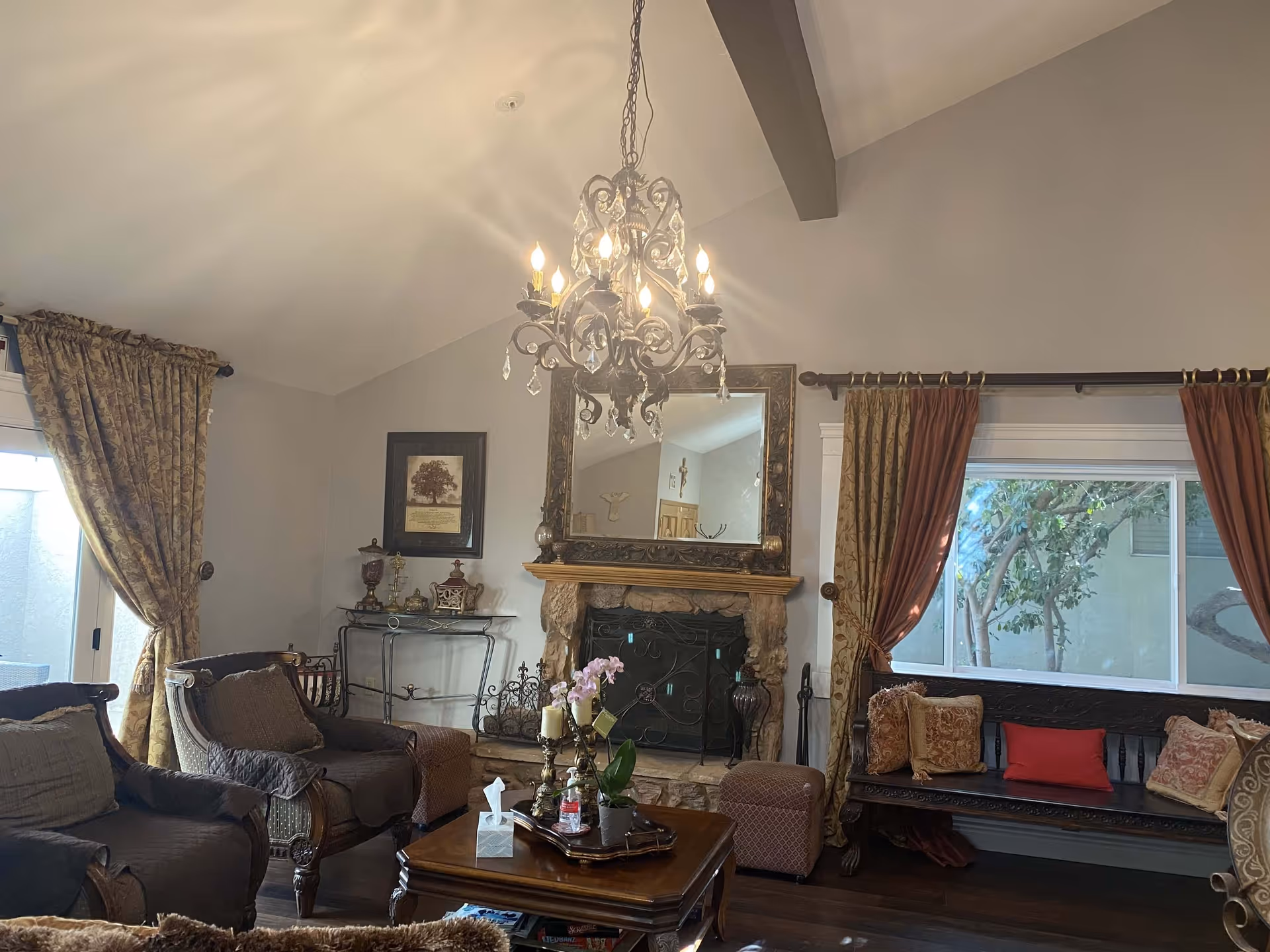 A cozy living room with a stone fireplace, an ornate mirror above it, and a decorative chandelier hanging from the ceiling. The room features comfortable armchairs, a wooden coffee table with a tissue box and plants, and a wooden bench with multiple cushions. Large windows with patterned curtains allow natural light to enter the space.