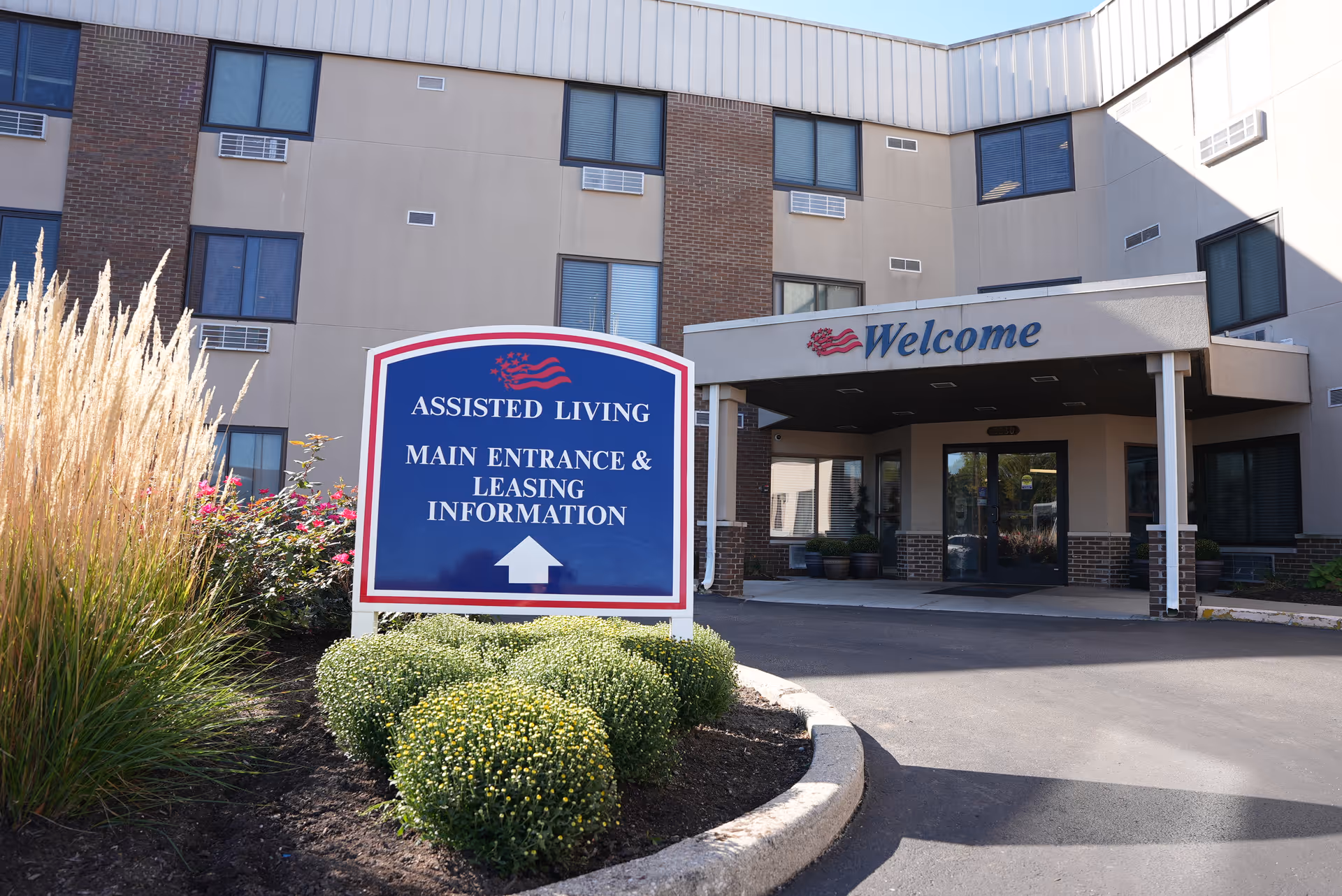 Exterior view of the entrance to an assisted living facility with a sign in front that reads 'Assisted Living Main Entrance & Leasing Information' and a covered entrance with the word 'Welcome' above the doors.