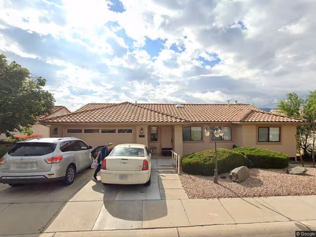 Single-story residential building with a tiled roof, two cars parked in the driveway, a person standing near one of the cars, and a landscaped front yard with bushes and rocks under a partly cloudy sky.