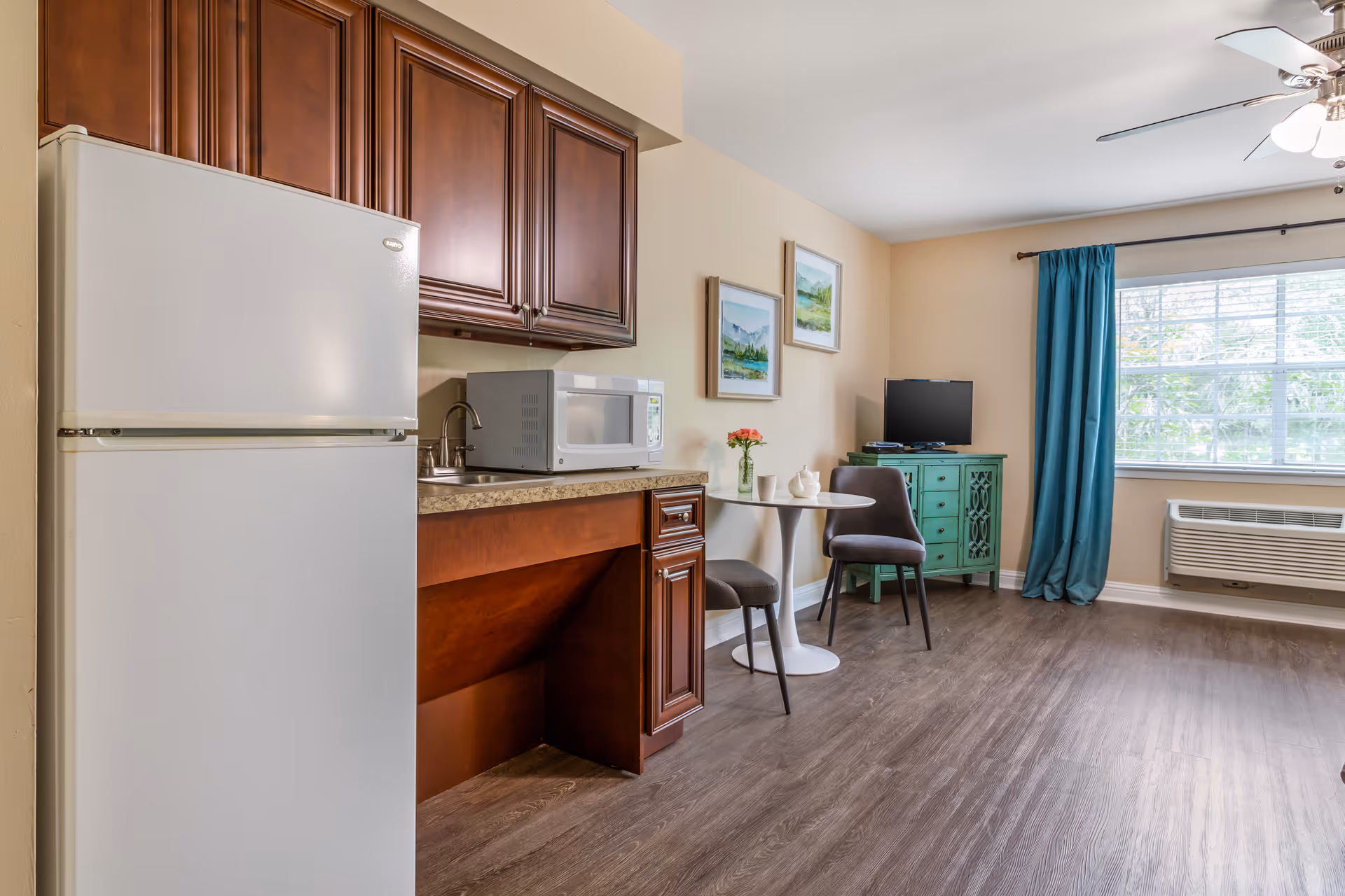 Studio-style living area with a kitchenette (refrigerator, microwave, sink), small round table with two chairs, TV on a teal cabinet, and a window with teal curtains.