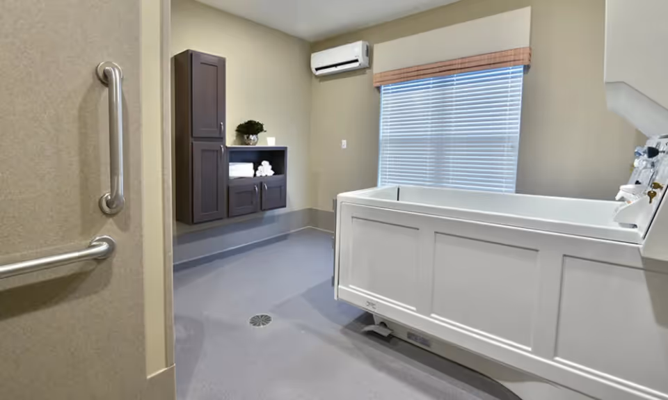 A clean and modern bathing room with a large white assisted bathing tub, beige walls, a window with closed blinds, a wall-mounted air conditioning unit, and dark wood cabinets holding towels and a small plant.