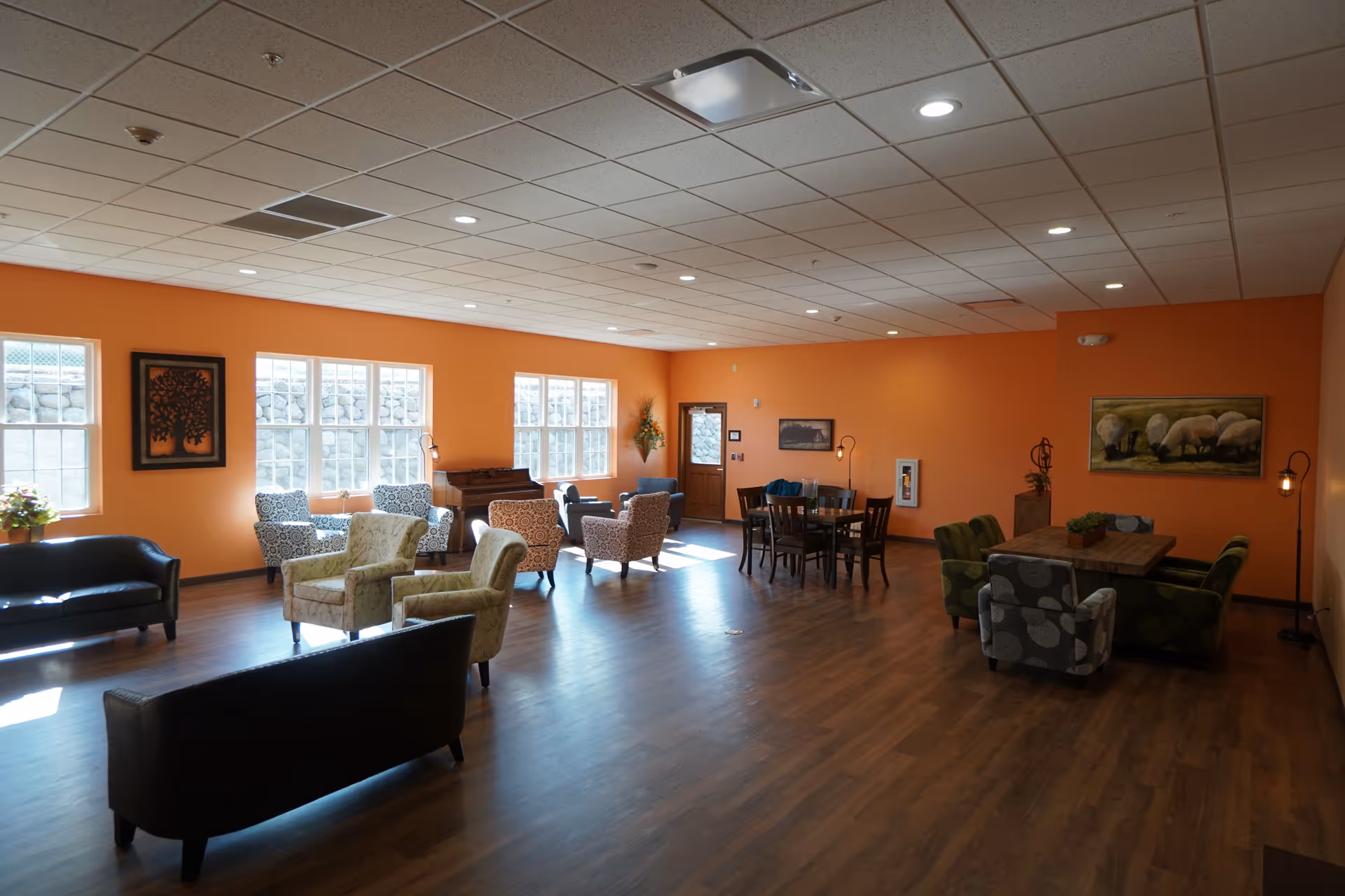 A spacious common area with orange walls and wood flooring, furnished with various chairs and sofas arranged in small groupings. There are multiple windows letting in natural light, framed artwork on the walls, and a piano near the windows. The ceiling has recessed lighting and a door is visible at the far end of the room.