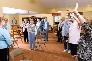 A group of elderly people standing in a spacious room with chairs arranged around them, participating in a group exercise or activity with their arms raised. The room has warm lighting, carpeted floors, and bookshelves in the background.