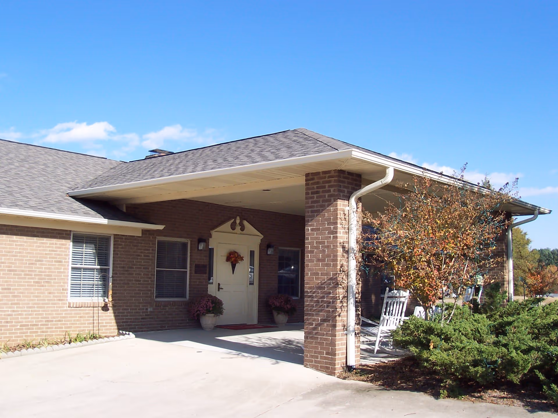 Exterior view of a brick building with a covered entrance supported by a brick pillar. The entrance door is white with a decorative wreath, flanked by two windows and potted plants. There are white rocking chairs on the porch next to a small tree with autumn-colored leaves. The sky is clear and blue.