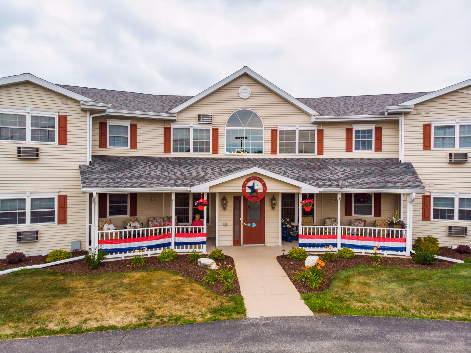 Front exterior view of a two-story senior living facility with beige siding and red shutters. The entrance features a covered porch decorated with red, white, and blue bunting and hanging flower baskets. There are several chairs on the porch with a few elderly people sitting and relaxing. The building is surrounded by a well-maintained lawn and flower beds.