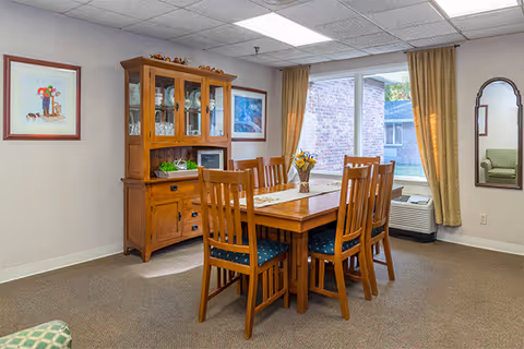 A dining room with a wooden table and six chairs with blue cushions. A wooden cabinet with glass doors displaying dishes is against the wall. There are two framed pictures on the walls, a window with beige curtains, and a mirror reflecting part of a green armchair. The room is carpeted and has a ceiling with fluorescent lights.