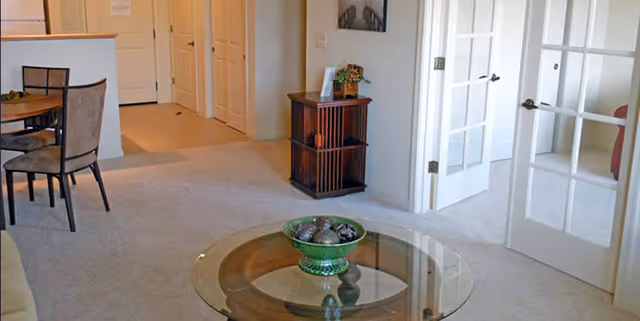 Carpeted living area with a glass coffee table holding a decorative bowl, a dining table to the left, and French doors opening to an adjacent room.