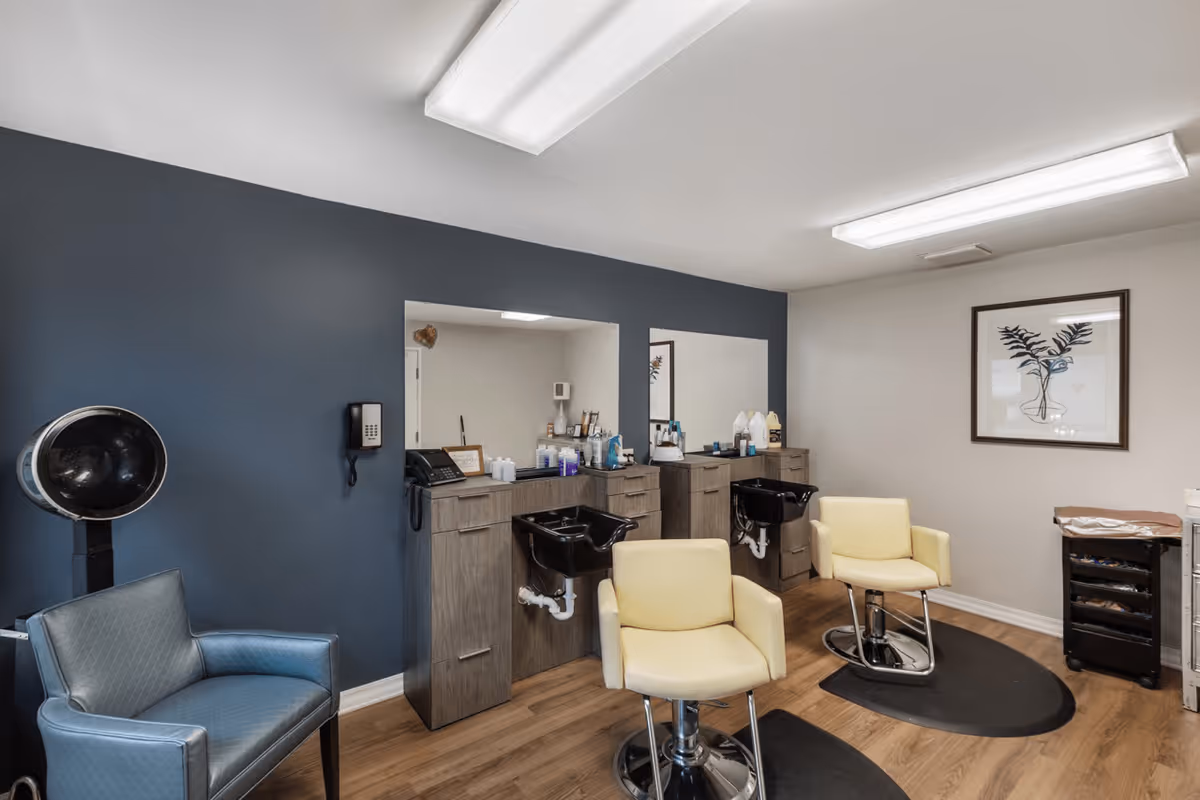 Interior view of a senior living facility's hair salon with two yellow salon chairs in front of two black hair washing sinks and mirrors. There is a blue wall with a mounted phone, a blue armchair with a hair dryer hood, wooden flooring, and a framed botanical artwork on the white wall.