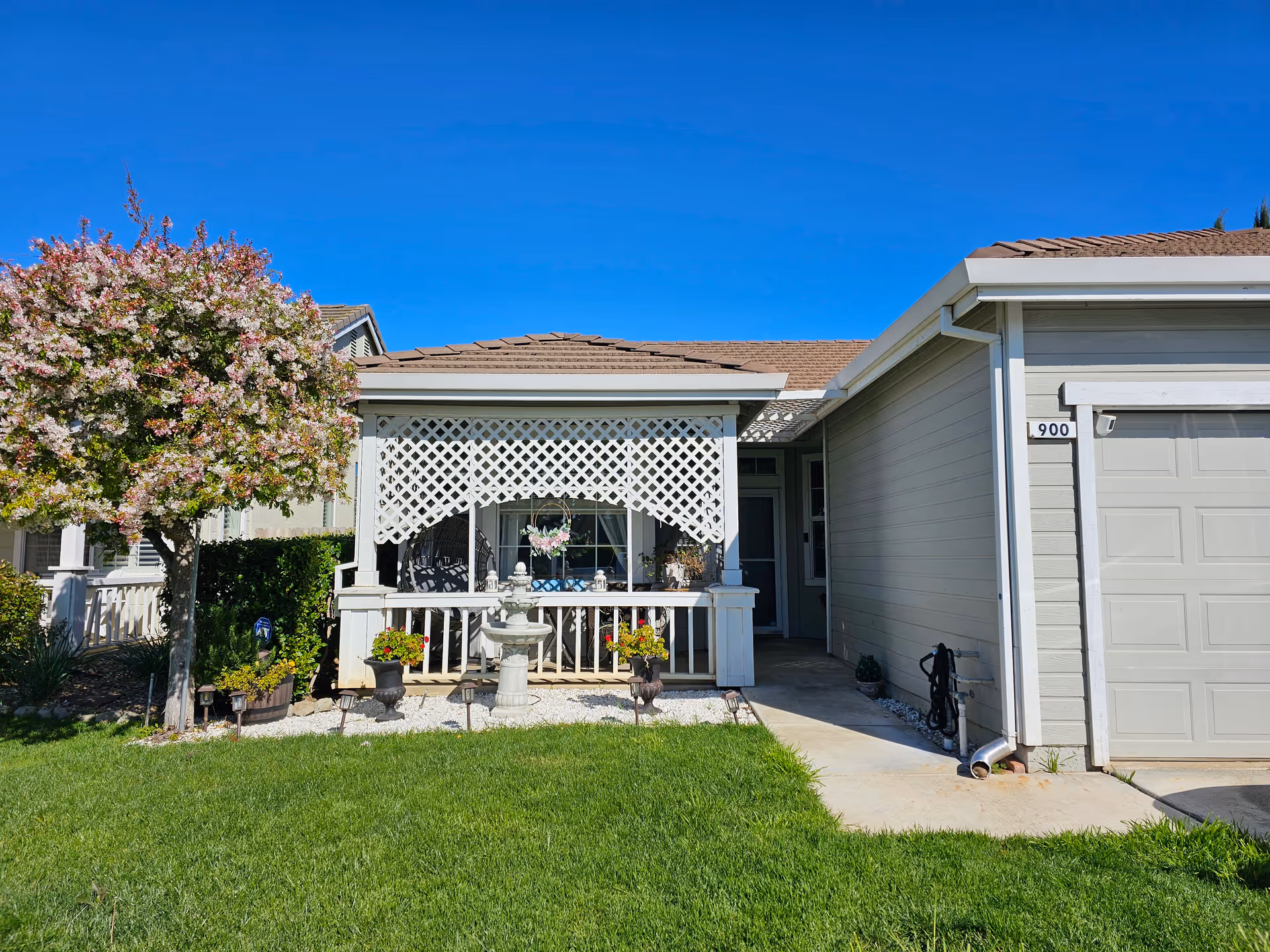 Front exterior view of a single-story house with a small porch featuring white latticework, a decorative fountain, potted plants, and a blooming tree on the left side. The house has light gray siding and a garage on the right side under a clear blue sky.