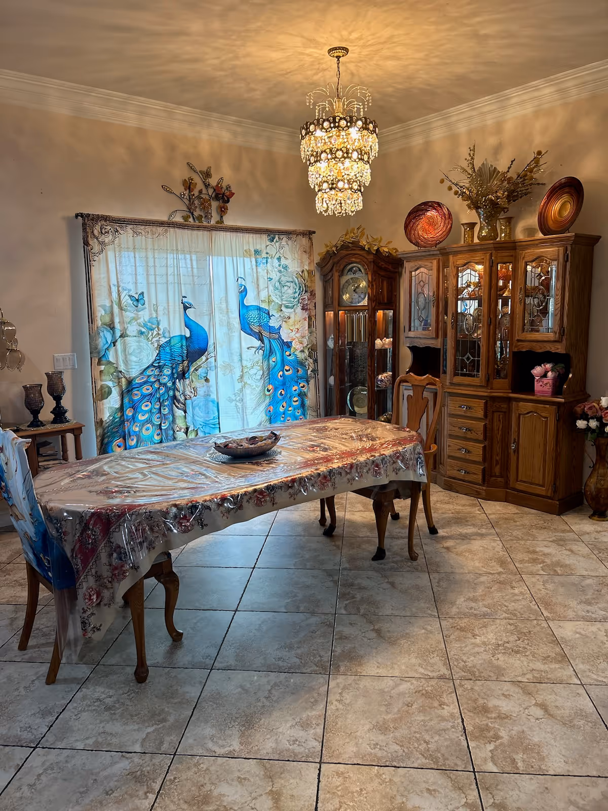 Dining room with a long table covered in plastic, peacock-themed curtains, a crystal chandelier, a grandfather clock and a wooden china cabinet.