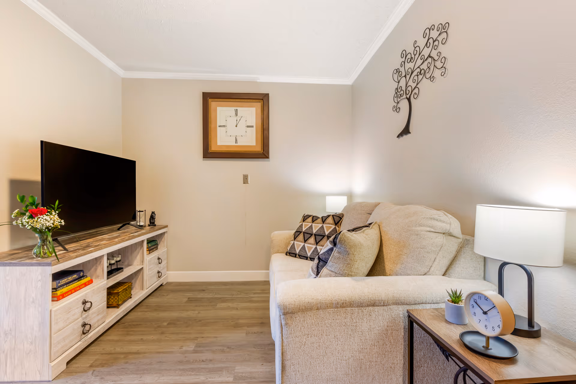 A cozy living room with a beige sofa adorned with patterned cushions, a wooden side table with a small plant, a clock, and a lamp. Across from the sofa is a wooden TV stand with a flat-screen television, books, and decorative items. The walls are light-colored with a framed clock and a metal tree wall decoration.