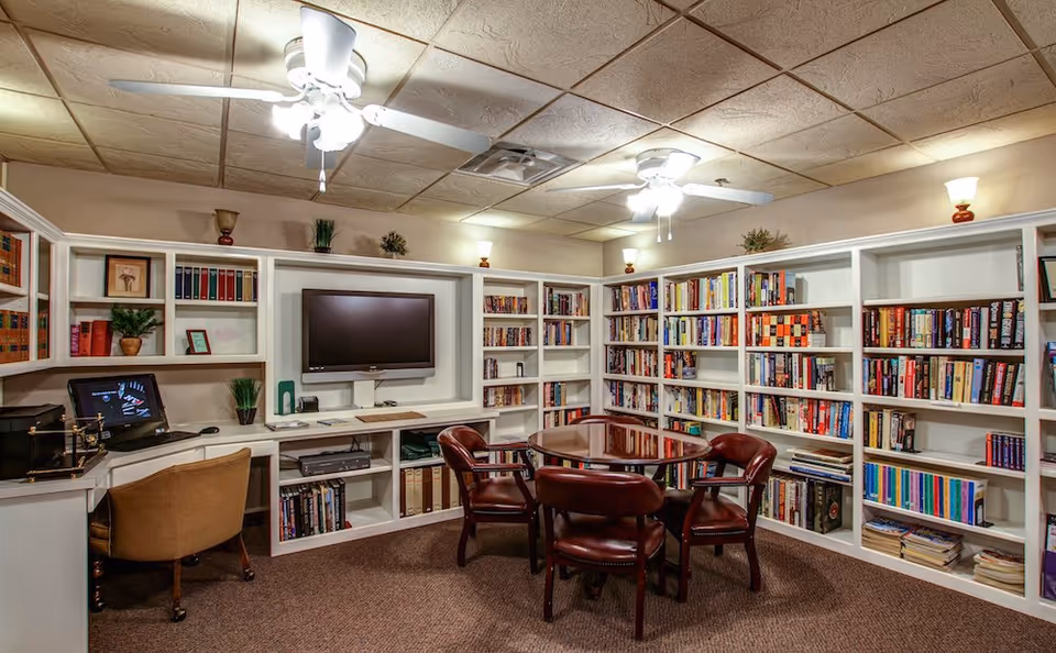 Cozy communal library/media room with built-in white bookshelves, a wall-mounted TV, a round table surrounded by leather chairs, a small desk area and ceiling fans.