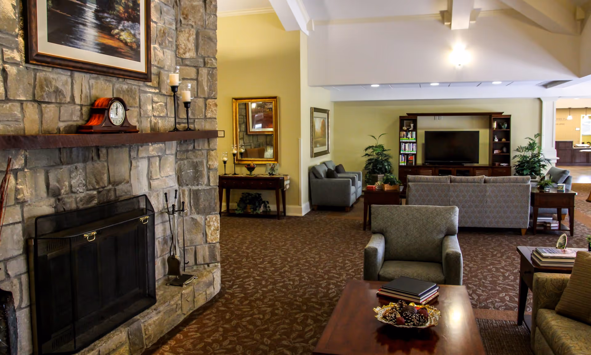 Lounge area with a stone fireplace, armchairs and sofas facing a TV cabinet in a senior living facility.