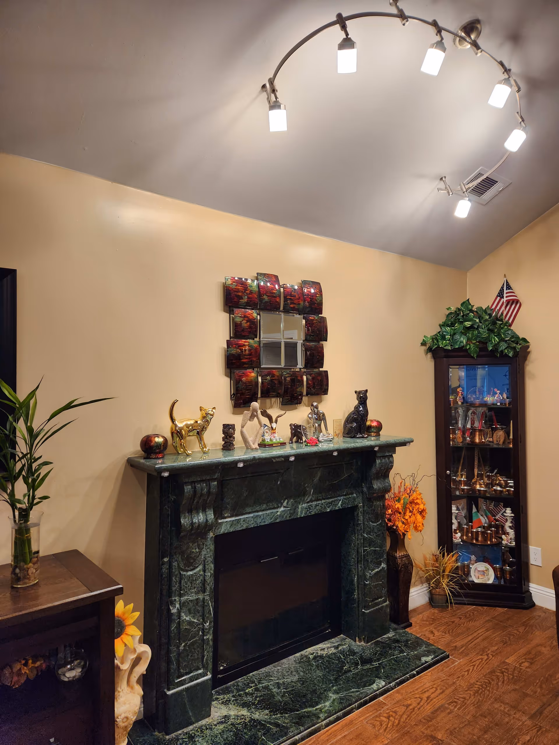 A cozy interior corner featuring a dark green marble fireplace mantel decorated with various small sculptures and figurines. Above the mantel is a decorative mirror with a red and black frame. To the right of the fireplace is a tall glass display cabinet filled with collectibles and topped with green foliage and a small American flag. To the left is a wooden side table with a plant in a glass vase. The walls are painted beige and the floor is wooden. A modern curved ceiling light fixture with multiple bulbs illuminates the room.