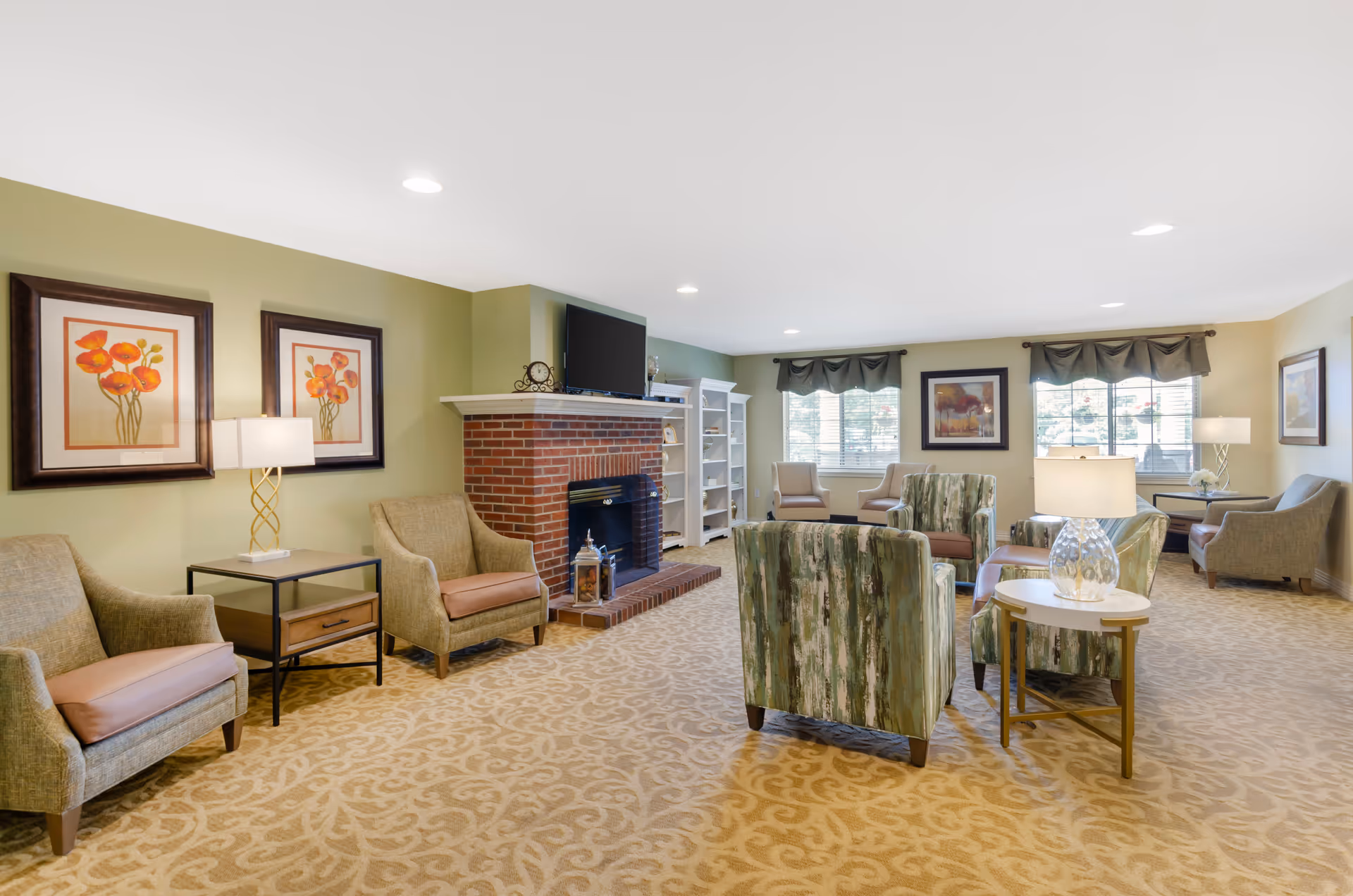 A spacious and well-lit living room with beige patterned carpet, green walls, and multiple seating areas including armchairs and sofas. There is a brick fireplace with a TV mounted above it, two framed floral paintings on the wall, and large windows with valances letting in natural light. Several table lamps provide additional lighting.