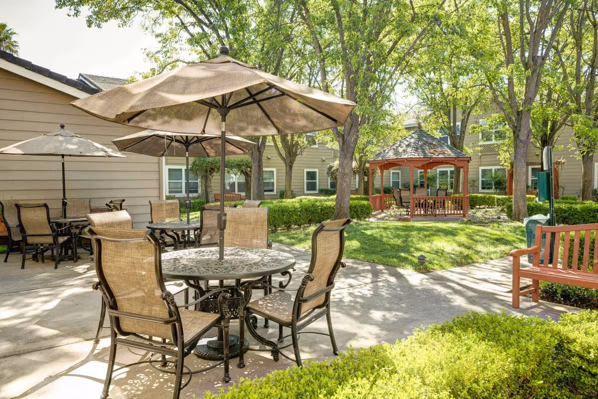 Sunny courtyard with patio tables and umbrellas, a gazebo, benches, and surrounding trees and apartments.