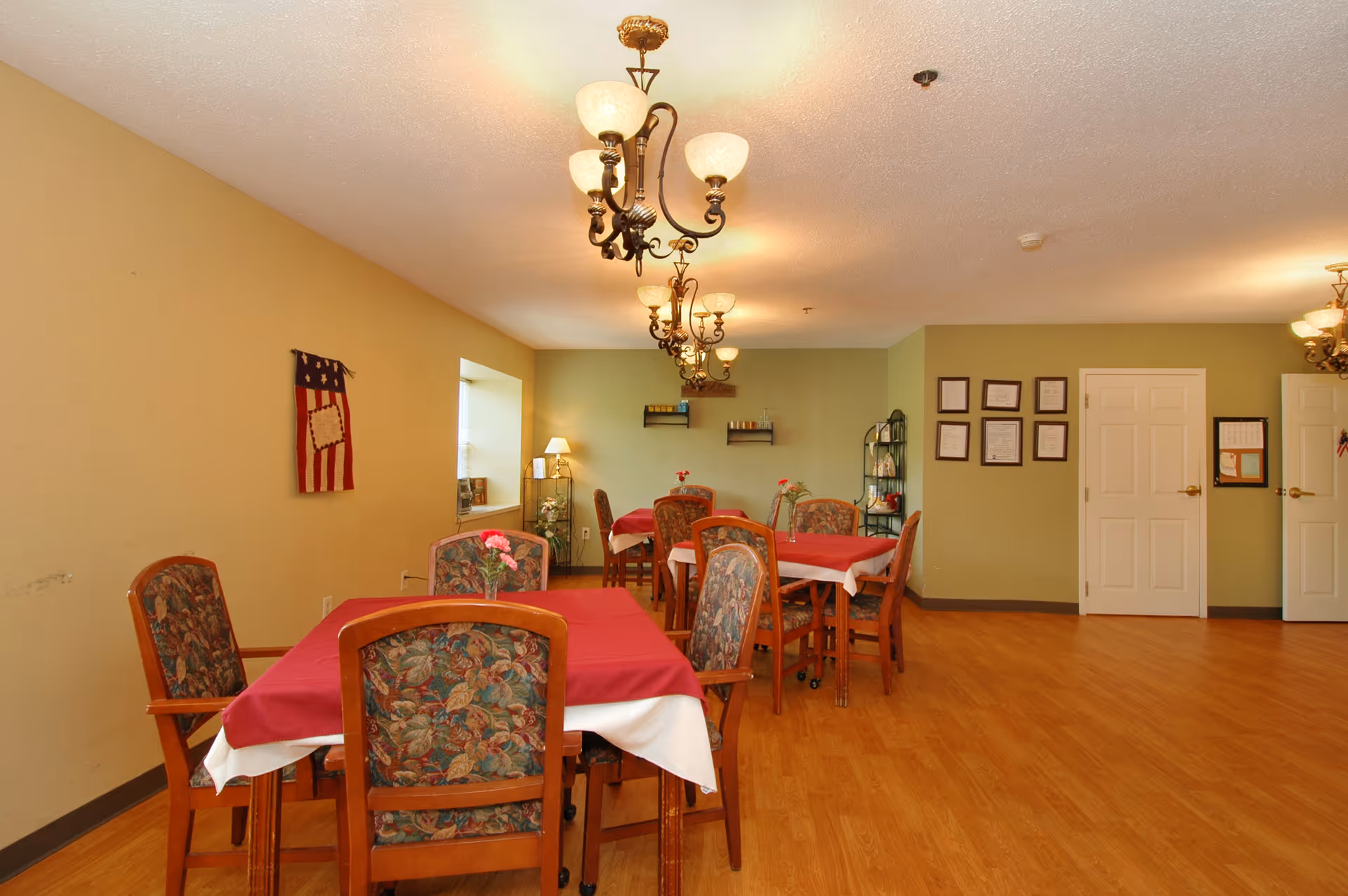 A dining room with several wooden tables covered with red and white tablecloths, surrounded by floral upholstered chairs. The room has wooden flooring, beige and light green walls, and is lit by ornate ceiling chandeliers. There are framed certificates on one wall and a small American flag decoration hanging on another wall.