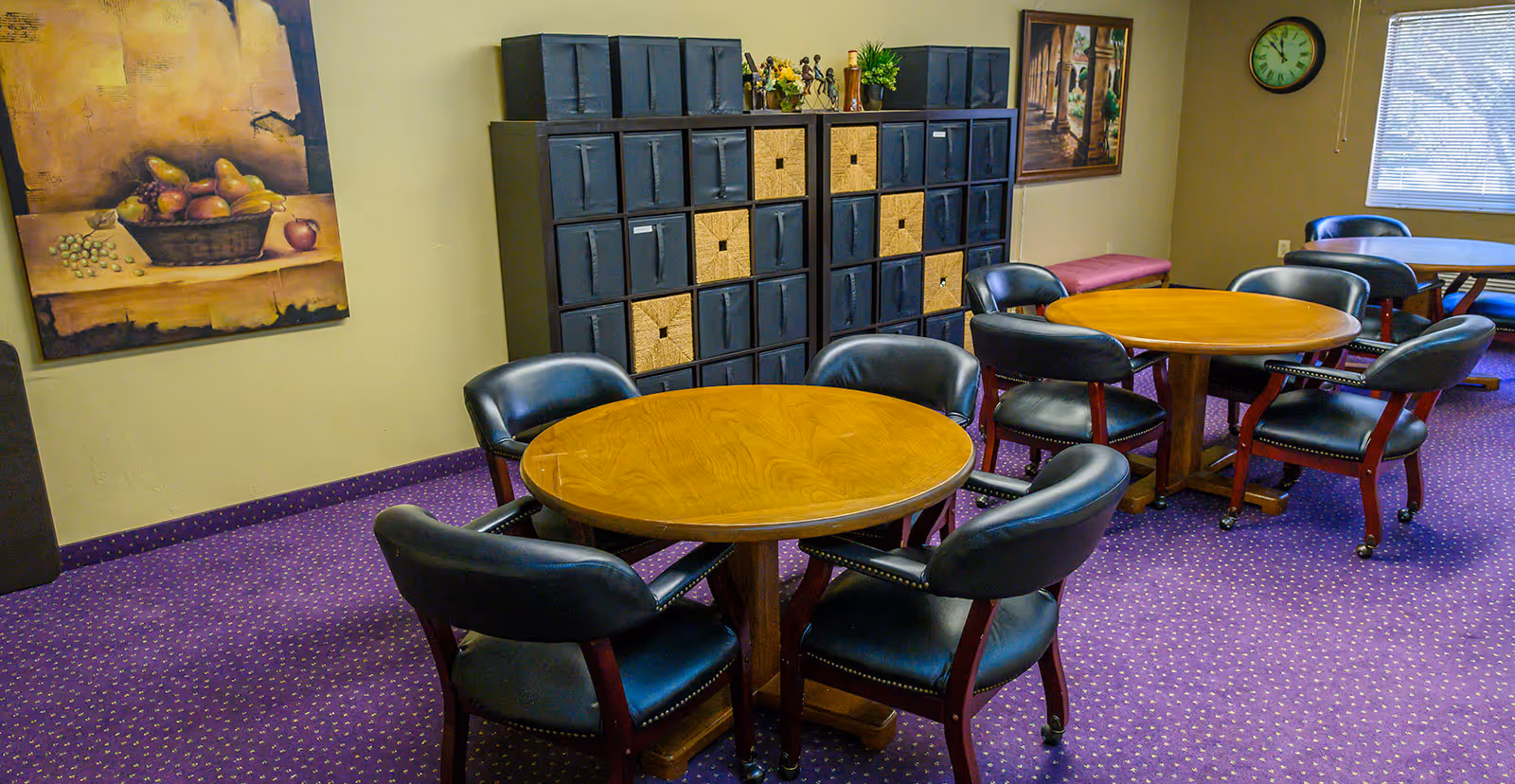 A communal room with round wooden tables and padded chairs, storage cubbies, wall art, and a purple carpeted floor.