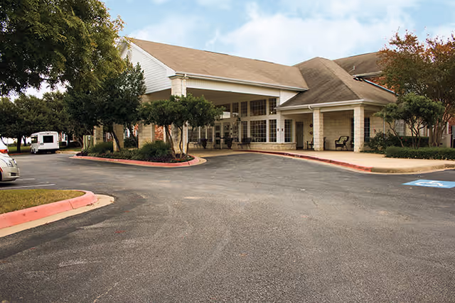 Front entrance and drive-up canopy of a single-story senior living facility with a paved parking area, landscaping, and covered porch seating.