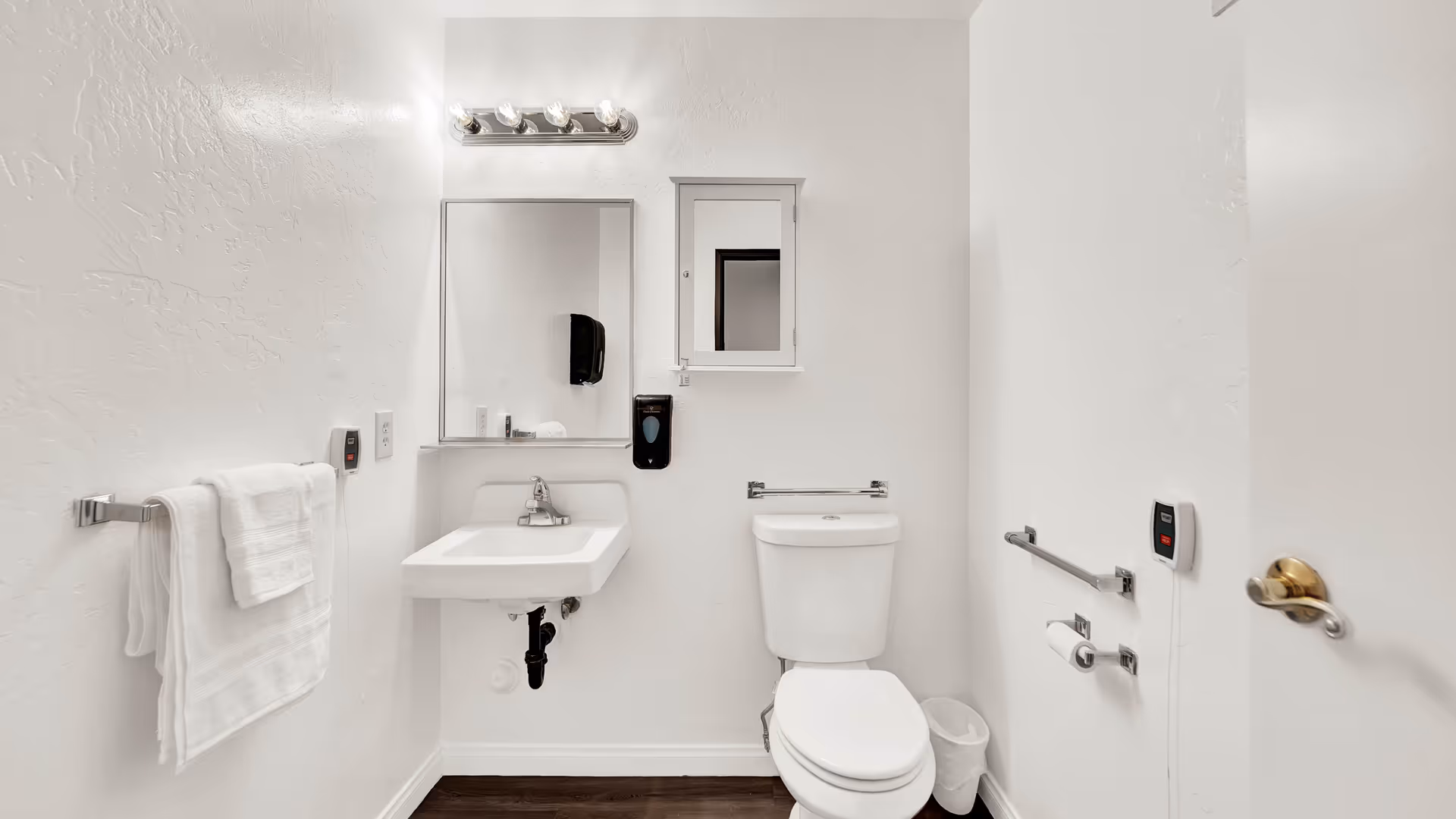 Small accessible bathroom with wall-mounted sink and mirror, toilet, grab bars, and towel rack.