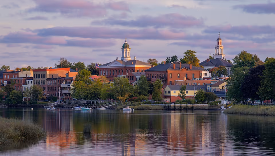View of a small town waterfront with historic brick buildings, trees, and boats docked along the shore under a partly cloudy sky during sunset.