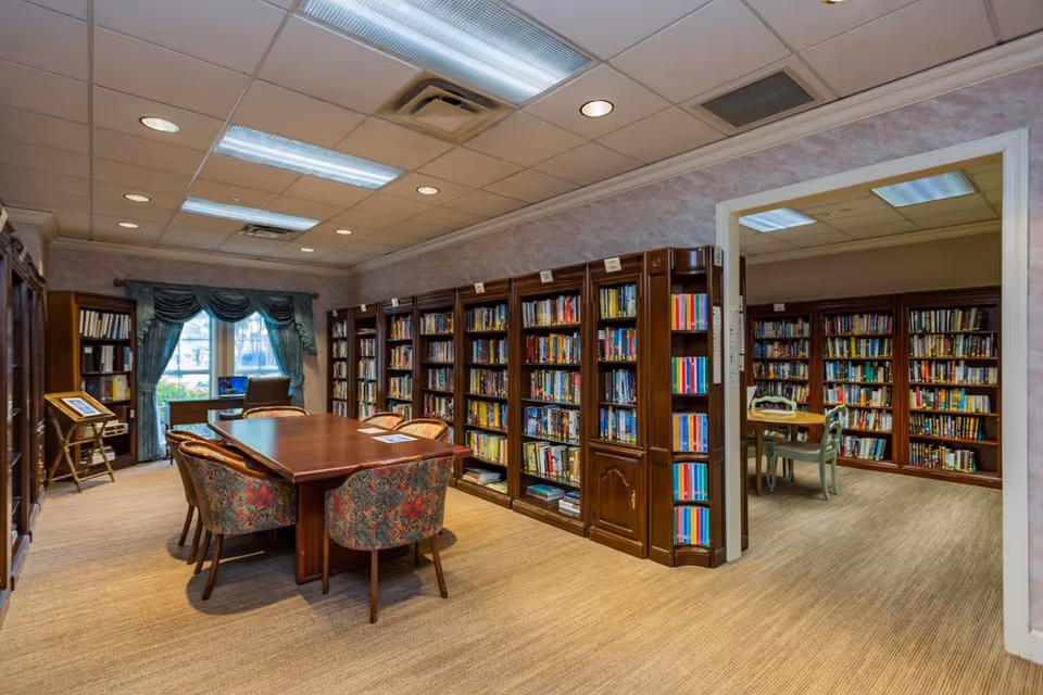 Interior view of a library room in a senior living facility with wooden bookshelves filled with books along the walls. A wooden table with six floral upholstered chairs is in the center of the room. There is a window with green curtains and a desk with a chair near it. Another room with more bookshelves and a small table with chairs is visible through an open doorway.