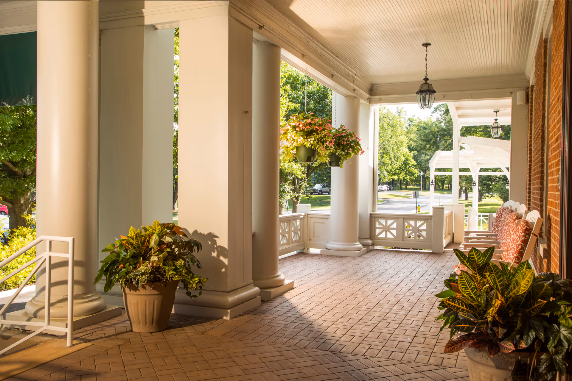 A spacious covered porch area with large white columns, hanging flower baskets, potted plants, and several cushioned rocking chairs along a brick wall. The porch overlooks a green outdoor area with trees and a street in the background.