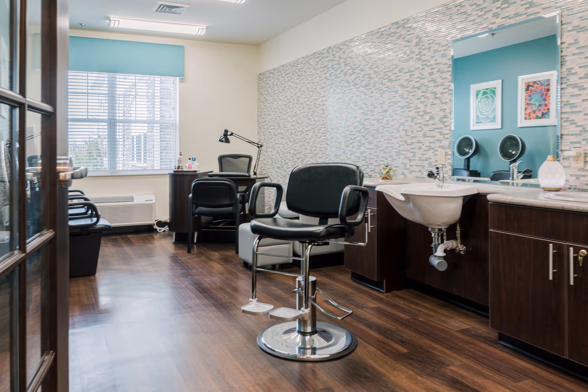A bright and clean salon room with a black salon chair in the center, a sink with a large mirror above it on the right, and a desk with a black office chair near a window with blinds in the background. The room has wood flooring and light-colored walls with a patterned accent wall behind the sink.