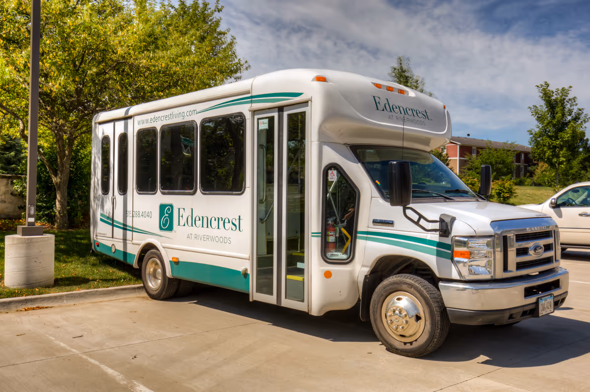 A white Edencrest at Riverwoods shuttle bus parked in a sunny parking lot with trees and buildings in the background.