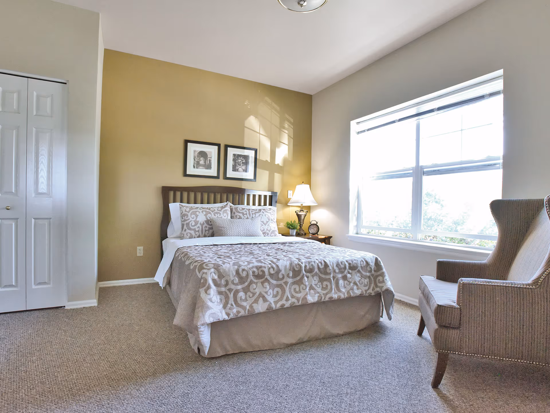 A bright bedroom with a bed covered in a beige and white patterned comforter and pillows. There is a wooden headboard, two framed pictures on the wall above the bed, a nightstand with a lamp, a small plant, and an alarm clock. A large window with blinds lets in natural light, and a cushioned armchair is placed near the window. The room has beige carpeting and light-colored walls.