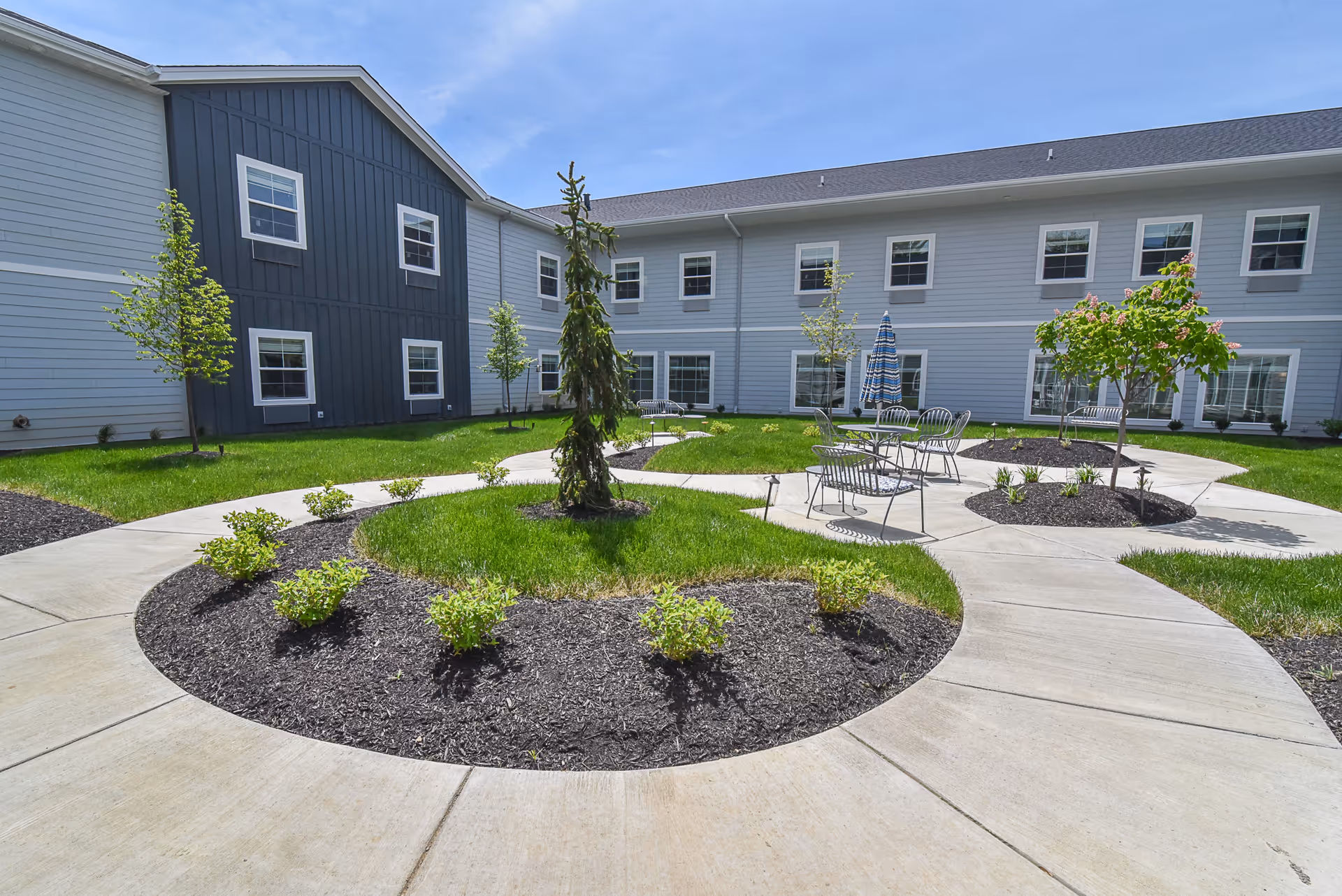 Outdoor courtyard area of a senior living facility with a circular concrete walkway surrounding landscaped garden beds with small trees and shrubs. There are metal chairs and tables with an umbrella for seating. The building with multiple windows surrounds the courtyard.