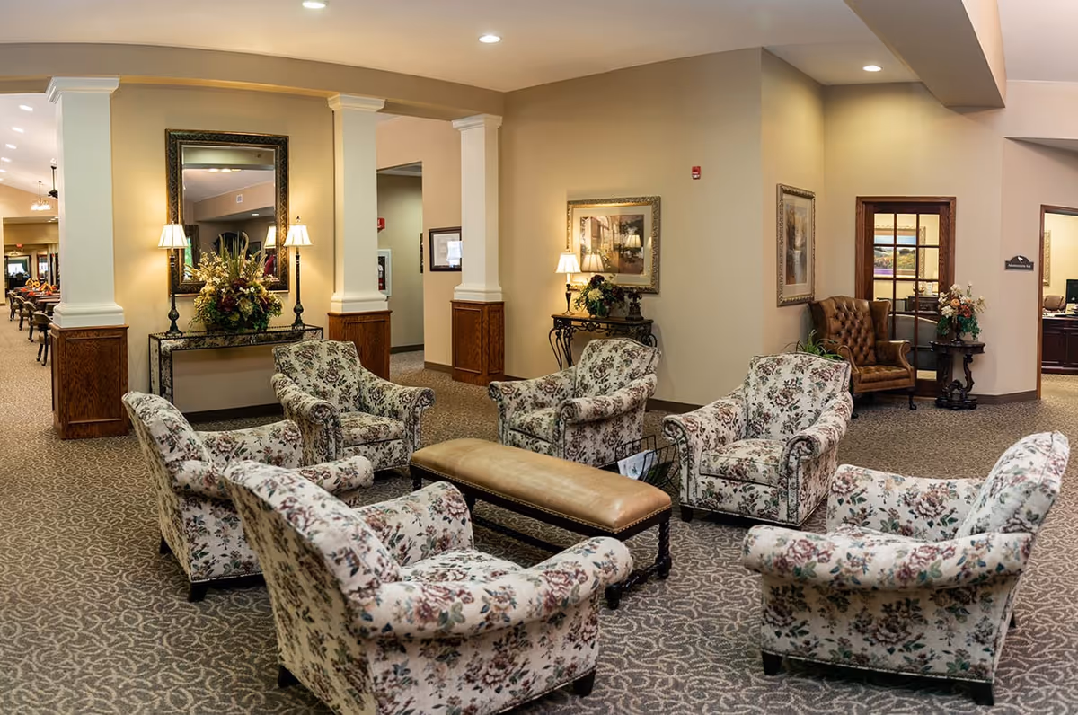 A cozy senior living facility common area with six floral upholstered armchairs arranged around a long cushioned bench. The room features beige walls, carpeted flooring with a patterned design, decorative columns, framed artwork, table lamps, and floral arrangements on tables and a console. A hallway and office area are visible in the background.