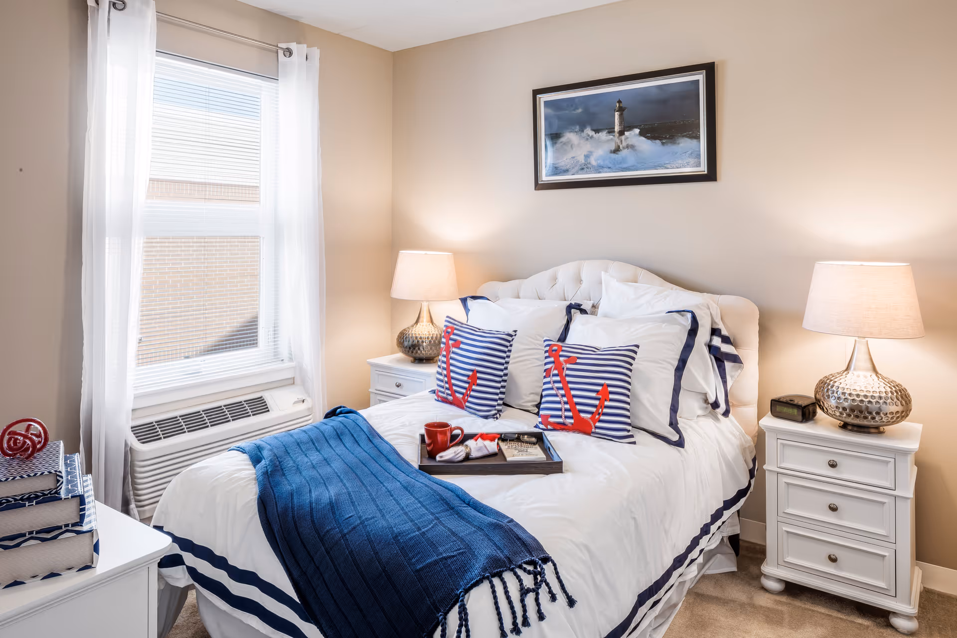 A cozy bedroom with a neatly made bed featuring white and navy bedding, two striped pillows with red anchors, and a navy throw blanket. There are two white nightstands on either side of the bed, each with a lamp. A window with white curtains is on the left wall, and a framed picture of a lighthouse and ocean waves hangs above the bed.