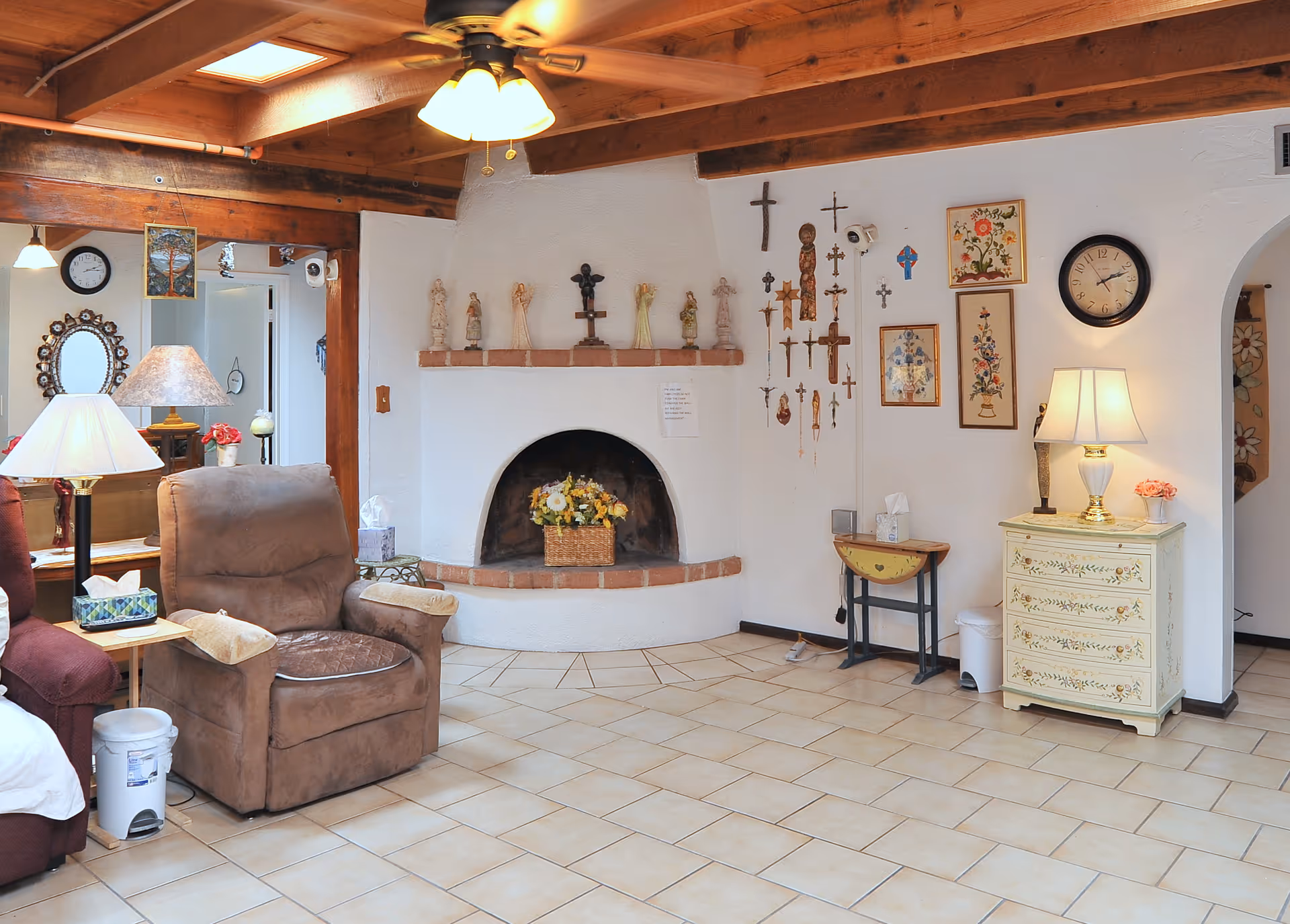 A cozy living room with a brown recliner chair, a red armchair, and a wooden side table with a lamp and tissue box. The room features a white stucco fireplace with a basket of flowers inside and various religious crosses and figurines displayed on the wall above and beside it. There is a ceiling fan with lights, a clock on the wall, and a small painted chest of drawers with a lamp and flowers on top. The floor is tiled, and wooden beams are visible on the ceiling.