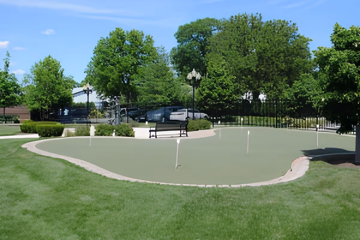 A landscaped outdoor putting green with flag markers, benches, and trees in the background.