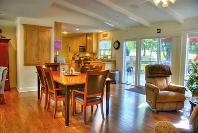 A bright and spacious common area featuring a wooden dining table with six chairs, a comfortable recliner chair near large glass sliding doors that open to an outdoor patio, and a kitchen area with wooden cabinets and appliances in the background.