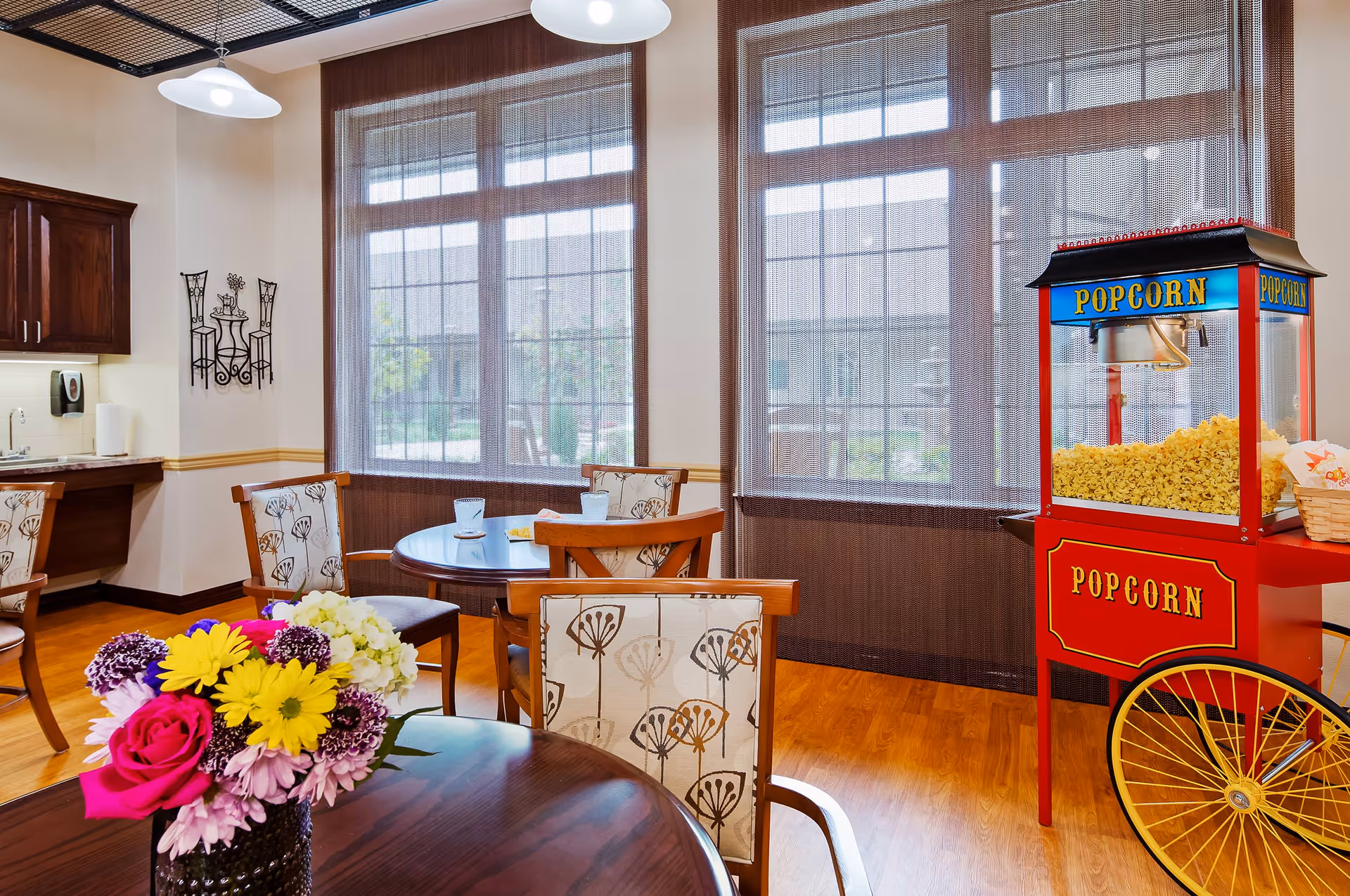 A bright dining area with wooden tables and chairs featuring floral upholstery. A colorful bouquet of flowers is on the foreground table. Large windows with sheer brown curtains allow natural light to fill the room. A red popcorn machine filled with popcorn is positioned near the windows. The room has wooden flooring and a small kitchenette area with cabinets and a sink in the background.