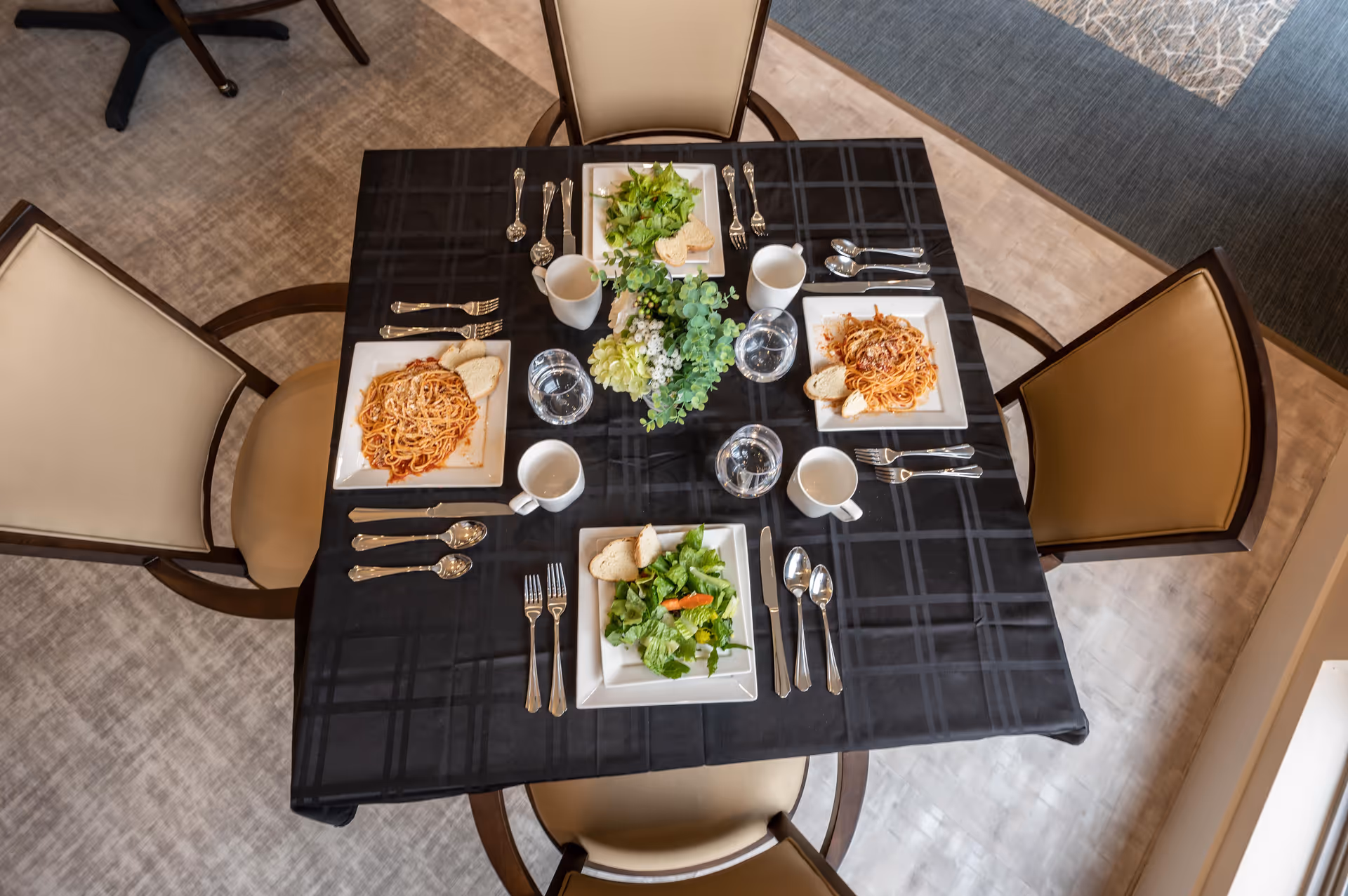 A dining table set for four with a black checkered tablecloth, each place setting includes a plate of food with spaghetti and bread or salad and bread, silverware, a white mug, and a glass of water. A floral centerpiece with green and white flowers is in the middle of the table. Four beige cushioned chairs surround the table.