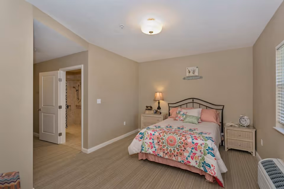 A bedroom with a bed covered in a colorful floral quilt and pink pillows. There are two nightstands on either side of the bed, one with a lamp and the other with an alarm clock. The room has beige walls, a window with blinds, and a door leading to a bathroom with a walk-in shower.