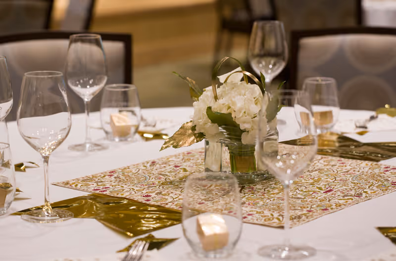 A close-up view of a dining table set with empty wine glasses, water glasses, and a floral centerpiece featuring white flowers and green leaves on a patterned table runner.